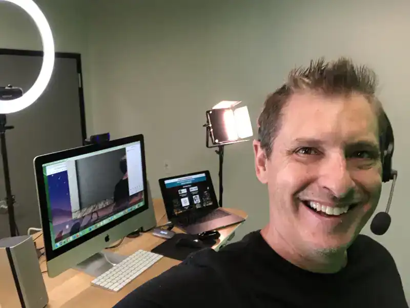 Scott Topper smiling in his hosting studio, wearing a headset, with a ring light and monitors behind him