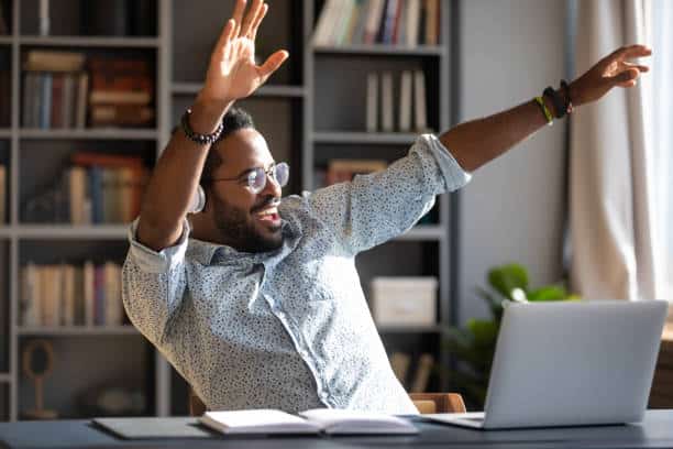 Overjoyed young African American man have fun dancing at workplace listening to music in headphones, happy biracial male worker or student enjoy good quality sound in modern wireless earphones